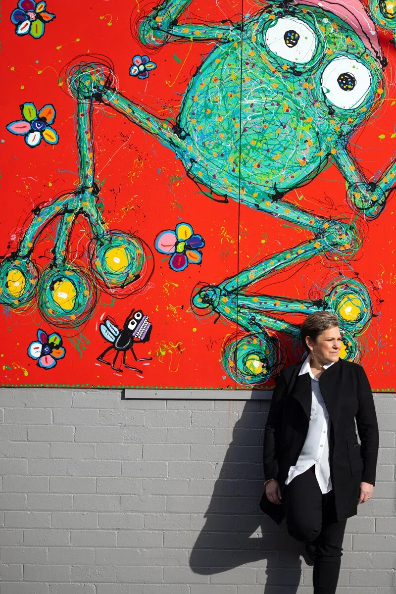 Sue leaning against a wall in front of a large, colourful frog mural by Anthony Breslin